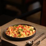 Colorful Indian chaat topped with crispy sev, pomegranate seeds, fresh coriander, and chutneys, served on a dark plate at a restaurant table.
