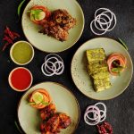 Overhead spread of Indian grilled specialties, including tandoori chicken, green-marinated paneer, and grilled meat kebabs, served with onion rings, lemon, mint chutney, and red sauce.