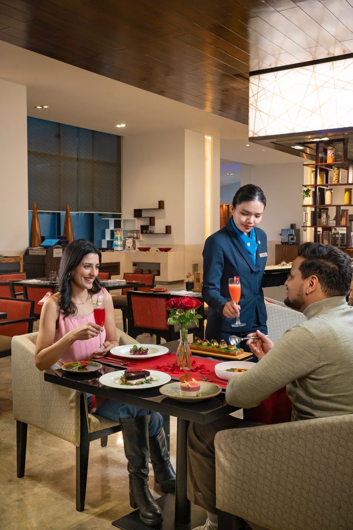 Couple enjoying a romantic dinner with plated desserts, roses and colorful drinks as a server presents a cocktail at Food Exchange Aerocity Delhi
