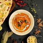 Overhead view of creamy Indian chicken curry garnished with cream and herbs, served with naan and surrounded by ginger, dried chillies and whole spices at Food Exchange Aerocity Delhi