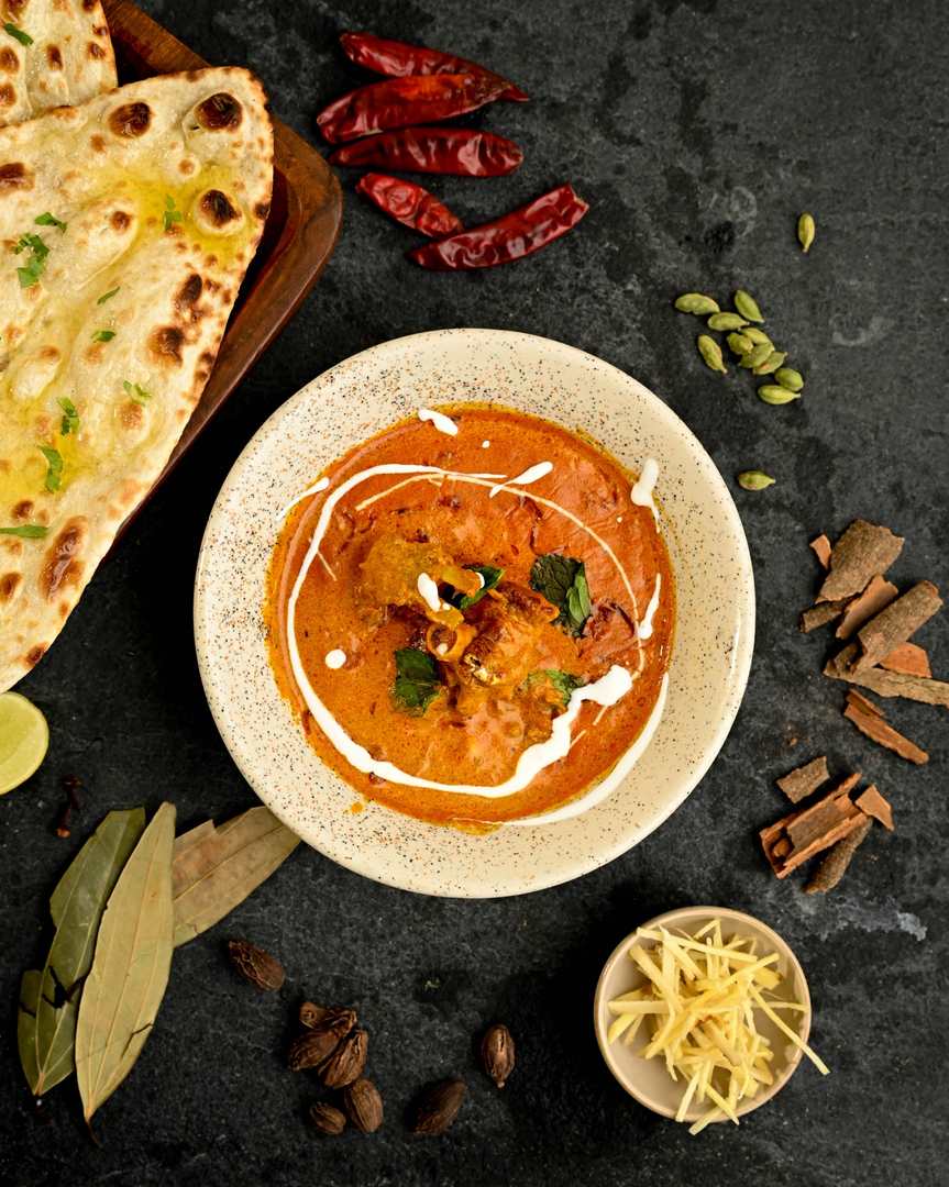Overhead view of creamy Indian chicken curry garnished with cream and herbs, served with naan and surrounded by ginger, dried chillies and whole spices at Food Exchange Aerocity Delhi