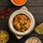 Overhead view of aromatic Indian biryani in a ceramic pot, served with raita, curry, ginger, dried chillies and whole spices at Food Exchange Aerocity Delhi