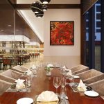 Elegant private dining area with a long wooden table set for guests, upholstered armchairs, modern pendant lights, abstract artwork, and views into a spacious contemporary restaurant.